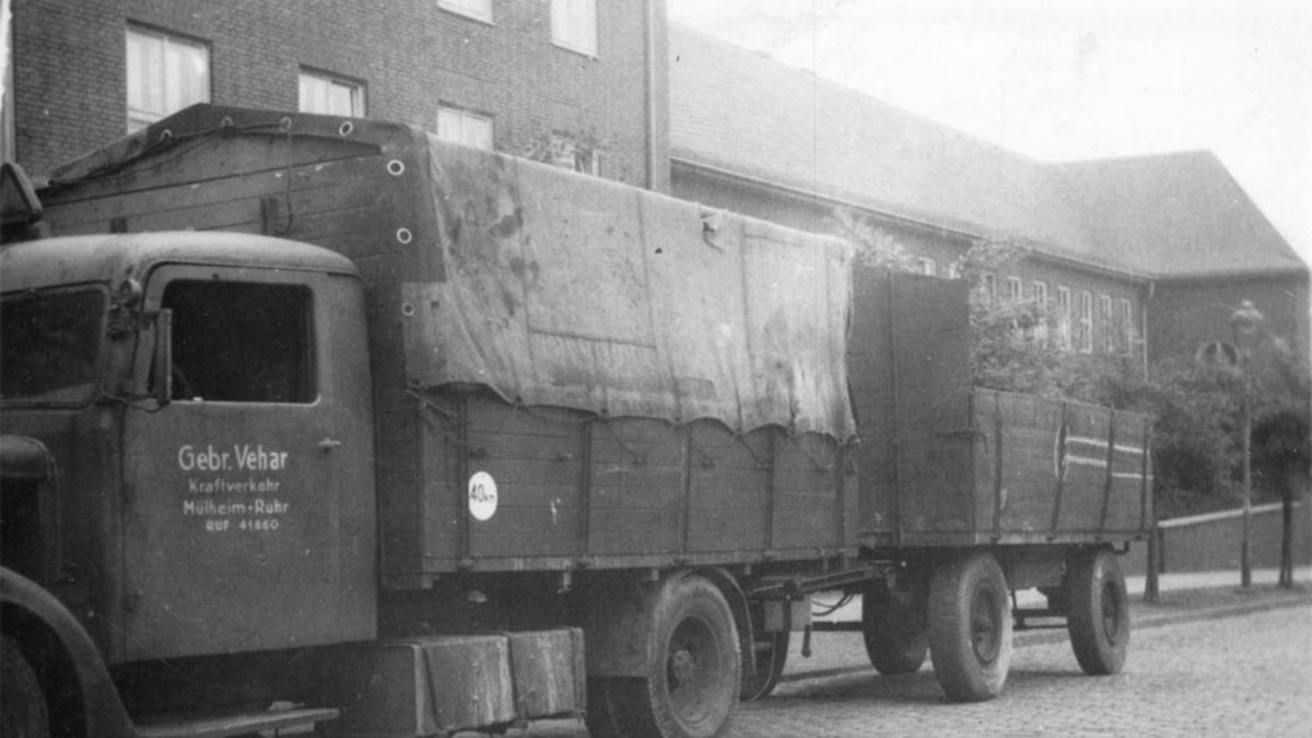 Schwarzweißfoto eines historischen LKW der Firma Gebr. Vehar vor einem Backsteingebäude, beladen mit einer Plane abgedeckter Fracht.